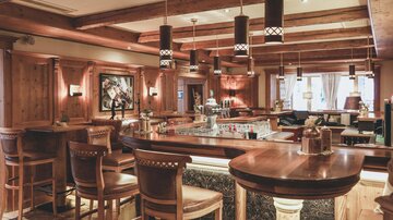 Eine gemütliche Bar mit Holzverkleidung und stilvollen Hockern. An der Theke stehen verschiedene Getränkeflaschen und es gibt sanfte Beleuchtung.