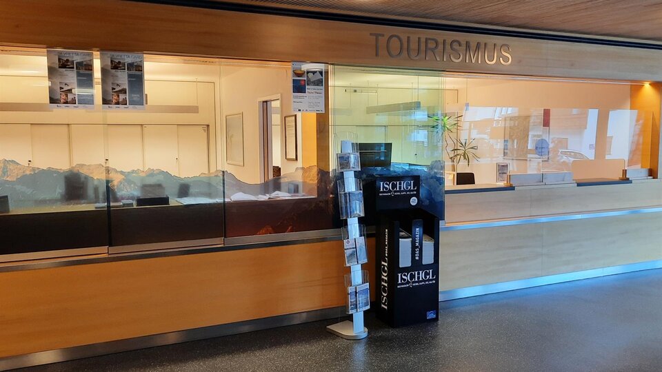 Ein Empfangsbereich für Tourismus mit einer Glaswand. Auf dem Boden steht ein Informationsständer über die Region Ischgl.