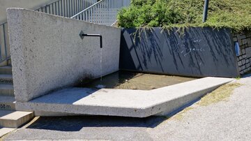 Ein moderner Brunnen aus Beton mit einem Wasserfluss und flachem Becken. Im Hintergrund sind grüne Sträucher zu sehen.