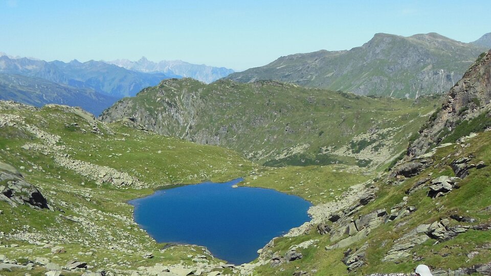 Ein schöner Bergsee umgeben von grünen Wiesen und Bergen. Der Himmel ist klar und blau.
