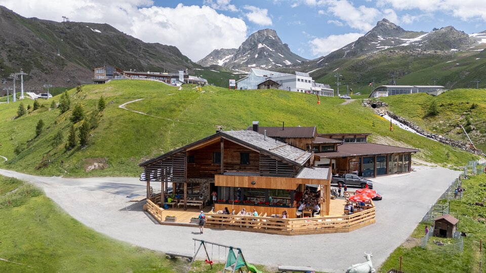 A modern wooden house in the mountains, surrounded by green meadows and rugged peaks. The sky is blue with some white clouds.
