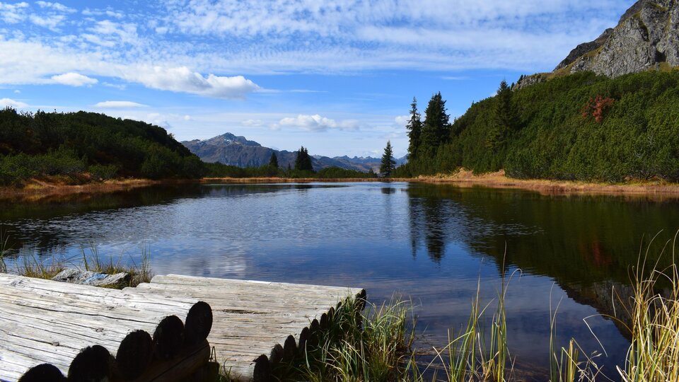 Ein ruhiger See umgeben von Bergen und Wald. Der Himmel ist klar mit einigen Wolken.