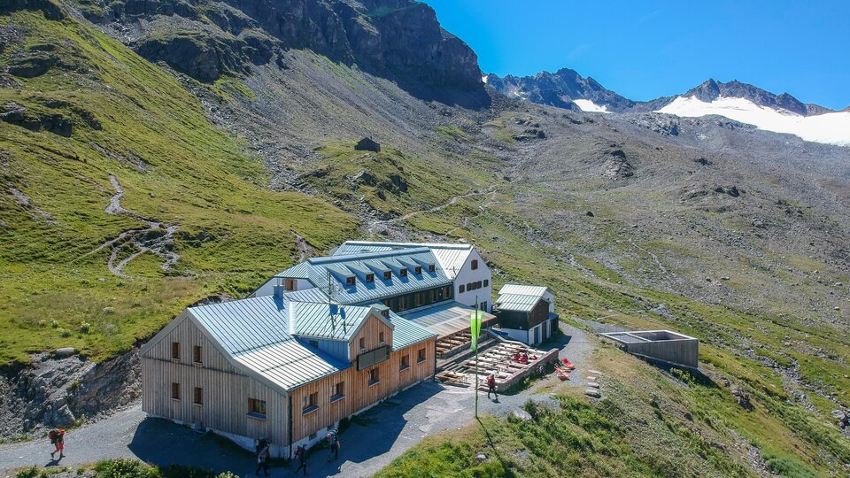 Eine Berghütte umgeben von grünen Wiesen und Bergen. Der Himmel ist klar und blau, ideal für einen Outdoor-Urlaub.