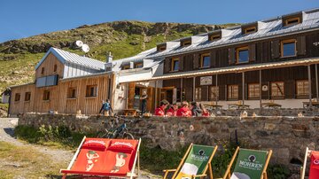 Ein gemütliches Berghaus mit Holzfassade und einer Terrasse. Im Vordergrund stehen bunte Liegestühle und mehrere Personen genießen die Aussicht.
