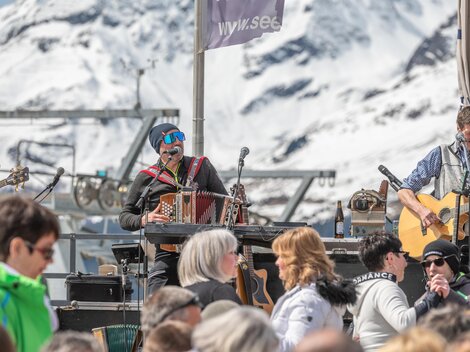 Eine lebhafte Musikveranstaltung in den Bergen mit schneebedeckten Gipfeln im Hintergrund. Verschiedene Musiker spielen vor einem Publikum in der Sonne.