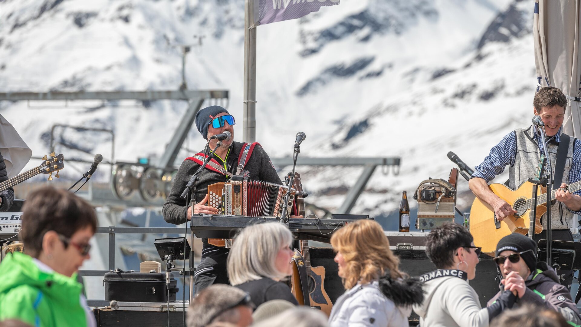 Eine lebhafte Musikveranstaltung in den Bergen mit schneebedeckten Gipfeln im Hintergrund. Verschiedene Musiker spielen vor einem Publikum in der Sonne.