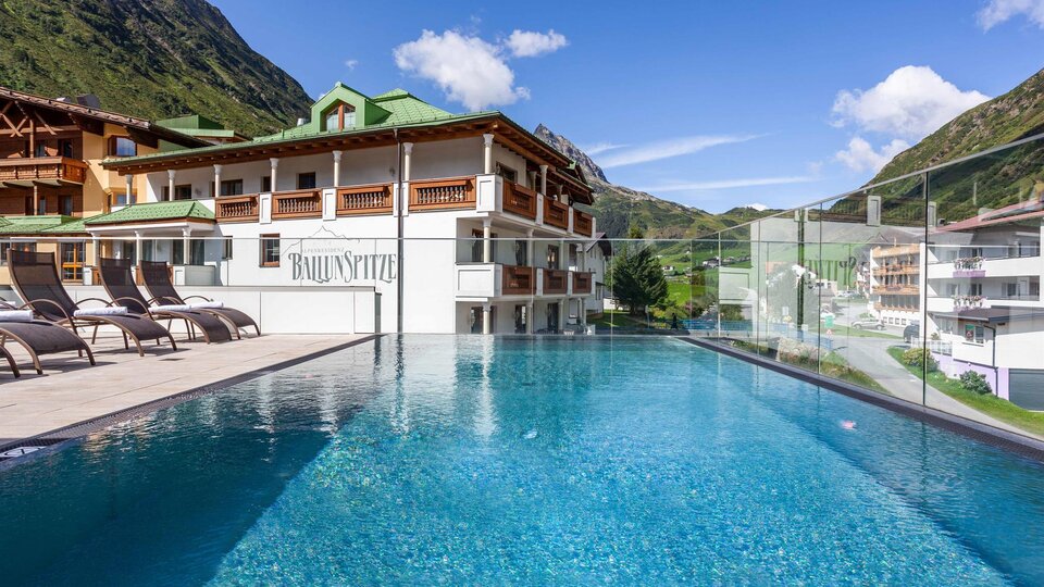 Ein modernes Hotel mit einem schönen Pool und Liegen. Im Hintergrund sind grüne Berge und ein klarer Himmel zu sehen.