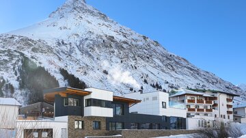Ein modernes Gebäude im Vordergrund, umgeben von schneebedeckten Bergen. Der klare, blaue Himmel vervollständigt die winterliche Landschaft.