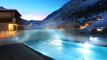 Ein schöner Außenpool in den Bergen bei Dämmerung. Die Lichter reflektieren auf dem Wasser und die schneebedeckten Berge sind im Hintergrund sichtbar.