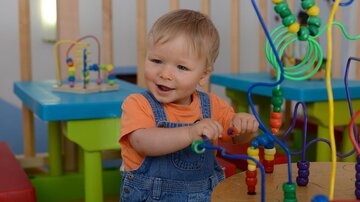 Ein kleines Kind mit blonden Haaren spielt an einem bunten Spieltisch. Es lächelt und bewegt die Perlen auf einem Strukturspielzeug.