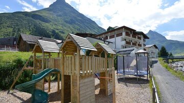 Ein Spielplatz aus Holz mit Rutsche und Trampolin in einer malerischen Berglandschaft. Im Hintergrund ist ein Hotel und hohe Berge zu sehen.
