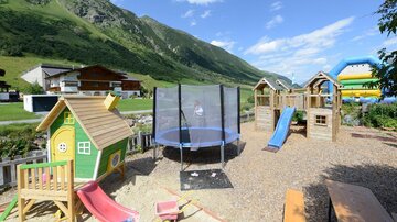 Ein schöner Spielplatz mit einem grünen Spielhaus, einer Rutsche, einem Trampolin und einem Hüpfburg im Hintergrund. Die Umgebung ist grün und bergig unter einem blauen Himmel.