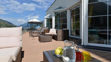 Ein einladender Balkon mit gemütlichen Möbeln und einem Tisch, auf dem ein Krug Wasser und Äpfel stehen. Im Hintergrund sind Berglandschaften und ein klarer Himmel sichtbar.