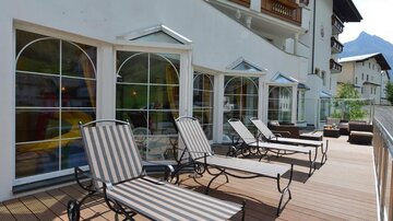 Eine Terrasse mit Liegen und großen Fenstern eines Hauses. Im Hintergrund sind Berge zu sehen.
