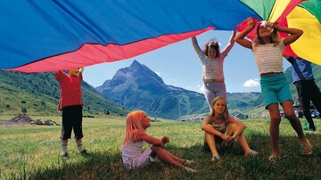 Eine Gruppe von Kindern spielt unter einem bunten Tuch in der Natur. Im Hintergrund sind grüne Wiesen und Berge zu sehen.