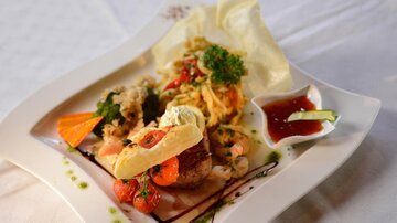 Ein kunstvoll angerichteter Teller mit verschiedenen Gerichten, darunter Fisch, Garnelen und Gemüse. Dazu gibt es eine Sauce und frische Kräuter.