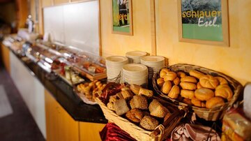 Ein reichhaltiges Frühstücksbuffet mit verschiedenen Brotsorten und Gebäck. Im Hintergrund sind zahlreiche Köstlichkeiten und Geschirr zu sehen.