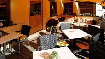 Ein gemütlicher Außenbereich mit Tischen und Stühlen in einem Restaurant. Die Wände sind in warmen Farben gestaltet, und es gibt bequeme Sitzgelegenheiten.