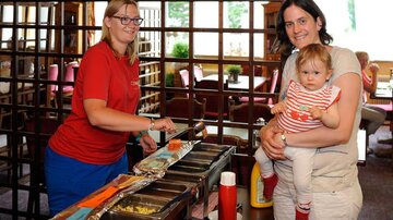 Eine Frau mit Brille serviert Hotdogs in einem Restaurant. Eine andere Frau hält ein kleines Kind auf ihrem Arm, während sie lächeln.