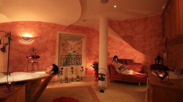 Ein entspannendes Badezimmer mit einer Badewanne und sanfter Beleuchtung. Eine Person entspannt im Bad, während eine andere auf einem Sofa liest.
