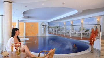 Ein eleganter Wellnessbereich mit einem Innenpool und Liegen. Eine Frau entspannt sich mit einem Getränk in der Hand.