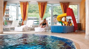 Ein Innenpool mit klarem Wasser und einer Rutsche für Kinder. Im Hintergrund sitzen Erwachsene auf Liegen und es gibt große Fenster mit Blick ins Grüne.