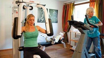 Eine Frau trainiert an einem Fitnessgerät, während eine andere Frau auf einem Laufband joggt. Im Hintergrund sieht man eine helle und freundliche Umgebung.