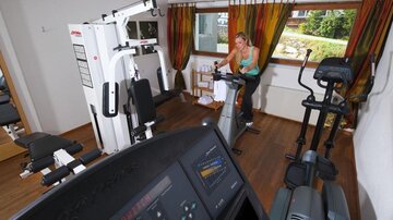 Ein gut ausgestatteter Fitnessraum mit verschiedenen Geräten. Eine Frau trainiert auf einem Ergometer.