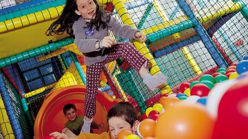Zwei Kinder spielen in einem bunten Spielparadies mit einer Rutsche und vielen Bällen. Ein Mädchen springt fröhlich in die Luft, während ein Junge im Hintergrund lachend im Bällebad spielt.