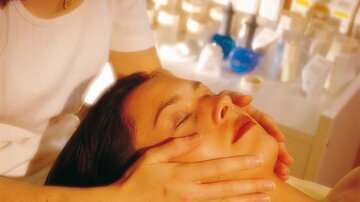 Eine Frau erhält eine Gesichtsmassage in einem Schönheitsstudio. Sie sieht entspannt aus, während eine andere Person ihre Hände sanft auf ihrem Gesicht platziert.