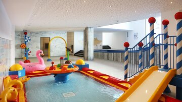 Ein farbenfroher, geschlossener Wasserbereich mit Rutschen und Spielgeräten für Kinder. Es gibt aufblasbare Spielsachen und ein glitzerndes Wasserbecken.
