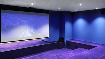 Ein moderner Heimkino-Raum mit einem großen Projektionsschirm und einer lila Teppichfläche. Die Wände sind in einem tiefen Blau gestrichen und die Decke hat dimmbare Lichter.