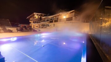 Ein beleuchteter Pool bei Nacht, umgeben von dampfender Atmosphäre. Im Hintergrund ist ein stilvolles Gebäude sichtbar.