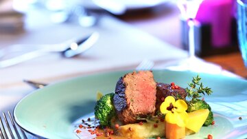 Ein kunstvoll angerichteter Teller mit zartem Fleisch, Brokkoli und buntem Gemüse. Die Präsentation ist elegant und ansprechend, mit einer essbaren Blume als Dekoration.