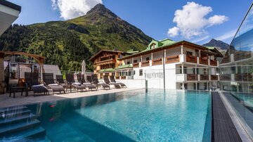 Ein modernes Hotel mit einem Swimmingpool und Liegen im Vordergrund. Im Hintergrund sind grüne Berge und ein blauer Himmel zu sehen.