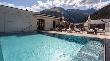 Ein schöner Pool mit klarem Wasser und Liegen. Im Hintergrund sind Berge und einen blauen Himmel zu sehen.