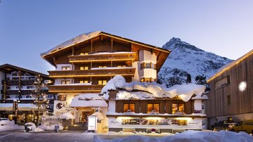 Ein charmantes Hotel im Schnee mit einem traditionellen Alpenstil. Im Hintergrund sind schneebedeckte Berge zu sehen.