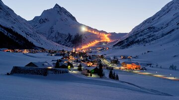 Eine verschneite Berglandschaft mit einem kleinen Dorf. Die Skilifte und Häuser sind sanft beleuchtet in der Dämmerung.