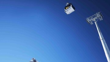 Eine Seilbahn schwebt über schneebedeckte Berge unter einem klaren blauen Himmel. In der Nähe steht ein großer Lichtmast.