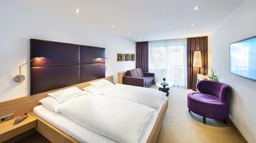 Ein modernes Hotelzimmer mit einem Doppelbett und einem gemütlichen Sofa. Große Fenster sorgen für viel Tageslicht und eine angenehme Atmosphäre. | © Rössle