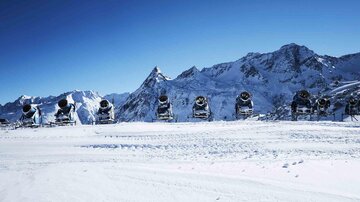Eine Schneelandschaft mit mehreren Schneekanonen, die auf einem klaren blauen Himmel stehen. Im Hintergrund sind majestätische Berge zu sehen.