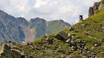 Ein Mountainbiker fährt einen steilen, felsigen Hang. Die Landschaft zeigt hohe Berge und eine grüne Wiese im Hintergrund.