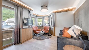Ein modernes Wohnzimmer mit großen Fenstern und Blick auf die Berge. Es gibt einen Fernseher, eine gemütliche Sofaecke und einen runden Tisch.