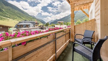Ein gemütlicher Balkon mit zwei Stühlen, umgeben von blühenden Blumen. Im Hintergrund sind grüne Berge und ein blauer Himmel mit schönen Wolken zu sehen.