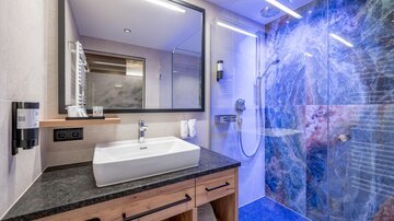 Ein modernes Badezimmer mit einer gläsernen Dusche und einem eleganten Waschtisch. Die Wand im Hintergrund hat ein kunstvolles, farbenfrohes Design.