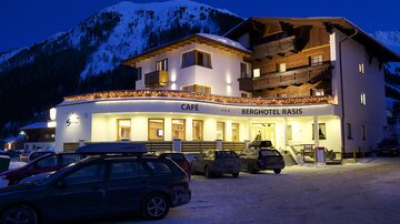 Ein gemütliches Berghotel mit einem Café, beleuchtet in der Dämmerung. Im Hintergrund sind schneebedeckte Berge sichtbar.