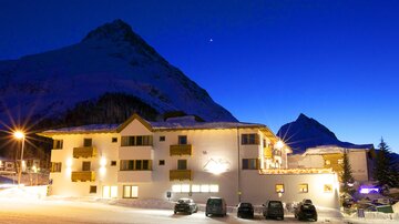 Ein gemütliches Hotel in einer verschneiten Berglandschaft bei Nacht. Der Himmel ist dunkelblau und die Berge sind majestätisch im Hintergrund sichtbar.