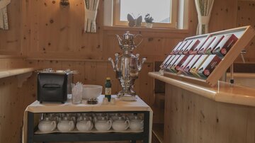 Ein Tisch mit einem Teekessel und verschiedenen Teesorten in einer Holzverkleidung. Im Hintergrund sind einige Fenster und eine gemütliche Atmosphäre sichtbar.