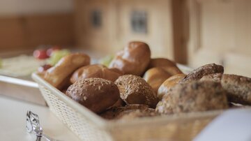 Ein Korb mit frischen Brötchen und Gebäck steht auf einer Holzablage. Im Hintergrund sind frische Zutaten und Küchenutensilien zu sehen.