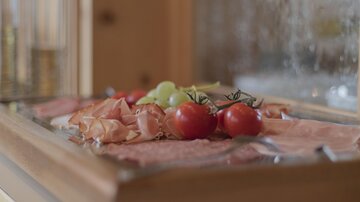 Ein von frischen Zutaten gefülltes Serviertablett mit Schinken, Tomaten und Trauben. Im Hintergrund ist eine holzverkleidete Wand zu sehen.